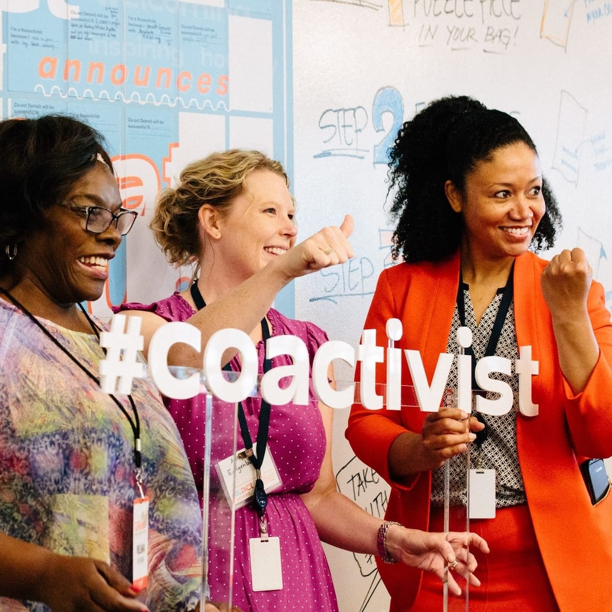 Three women standing next to each other and smiling in front of a logo that says "#coactivist" in white text color.